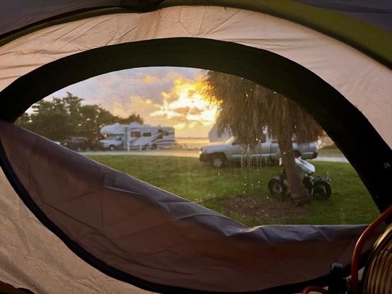 Picture of the sunset over the water on a rainy evening from inside our truck tent. Our e-bikes are outside, tied with our chains under a palm tree. Some RV campers can be seen farther rout, as well as a pickup truck behind the tree where our e-bikes are tied to.