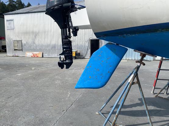 Photo of the stern of the sailboat, showing the black outboard motor and the blue rudder. The boat is on land, on jackstands.