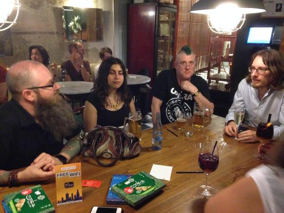 Four conference speakers sit around a wooden table in a dimly lit basement bar in Barcelona, mid-conversation over beers and sangria. A bearded, tattooed man on the left and a long-haired man on the right talk across the table while a long-haired Greek woman and a man with a green mohawk listen.