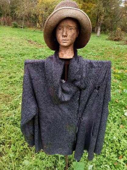 A plastic make-up head stuck on a steel traffic sign post wearing a hat and fur coat, overlooking a garden. The face is somewhat spotty black from some kind of plastic UV breakdown or maybe mould.