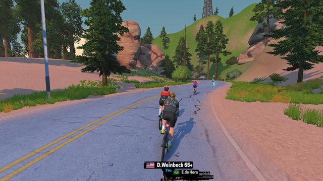 Screenshot aus Zwift: Mehrere Radfahrer fahren auf einer leicht ansteigenden, kurvigen Asphaltstraße durch eine Landschaft mit rötlichen Felsen, grünen Hügeln und hohen Nadelbäumen. Im Vordergrund fährt der eigene Avatar in schwarzer Radkleidung hinter anderen Fahrern. Die Straße führt in ein bewaldetes Tal.