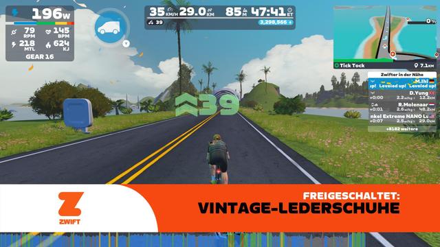 Screenshot aus Zwift: Der eigene Avatar fährt auf einer geraden Küstenstraße zwischen Palmen, grünen Hügeln und türkisblauem Wasser. Eine große grüne 39 zeigt den Aufstieg auf Level 39 an. Am unteren Bildrand erscheint ein orangefarbenes Banner mit der Meldung „Freigeschaltet: Vintage-Lederschuhe“. Oben sind 29,0 km Strecke, 85 Höhenmeter und 47:41 Minuten Fahrzeit zu sehen.