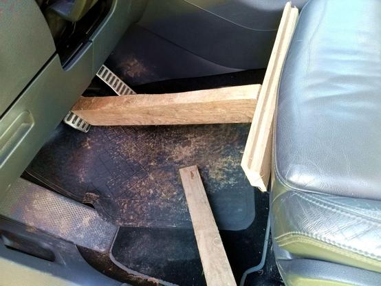 Two pieces of wood wedged between seat front and brake pedal, holding the pedal down. A spare piece that was the wrong length is also present. The rubber floor mat is dirty.