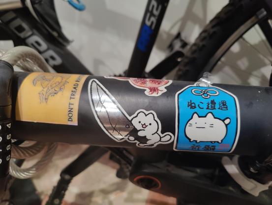 En el cuadro de la bici, una pegatina de un gusano de dune y dice "don't tread rhythmically", un gato adorable con un cuchillo, un amuleto japonés con un gato serio y parte de una granada