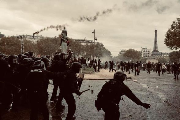 A gang of armored cops (flics) in riot gear, throwing tear gas into a mass of vulnerable protesters. They’re in a city square with a monument statue, under a cloudy sky, with the Eiffel Tower in the background.