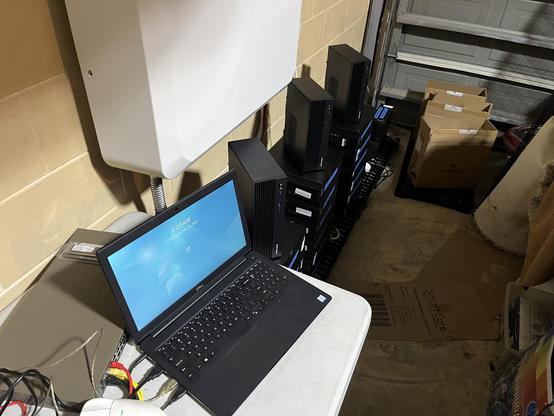 Cluttered garage with a laptop and network switch on a table in the foreground and stacks of computers at floor level in the background