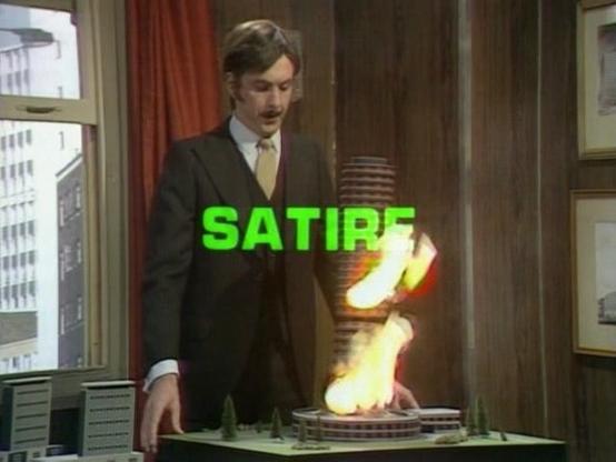Eric Idle dressed up like a businessman with a fake moustache presenting an architectural model of a proposed building that has caught on fire.

The word "satire" has flashed on screen in green as he looks at his burning sculpture keeping a deadpan expression

From monty python's flying circus, series 2, "The Buzz Aldrin Show."  The 17th episode overall.