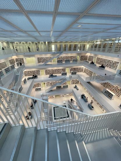 Foto des Innenraums der Stuttgarter Stadtbibliothek. Über 5 Etagen verbinden Treppen die quadratischen Etagen, an deren Außenwänden weiße Bücherregale stehen. Vor diesen Sitzmöbel auf denen Menschen in Bücher vertieft sind