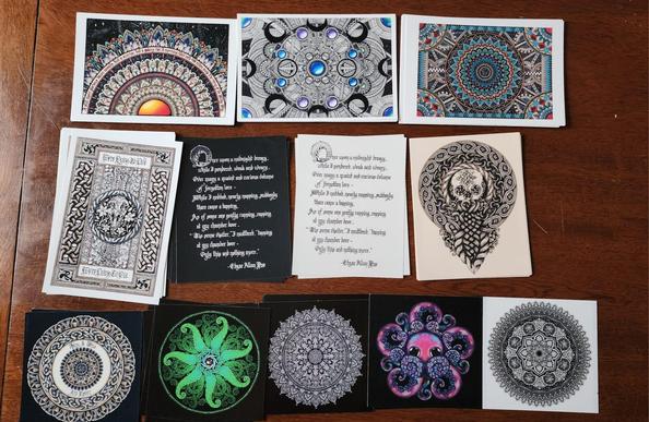 An assortment of prints on a table; some are postcard sized and some small squares. There are colourful mandalas, tentacles, black line art, calligraphy, Celtic knotwork