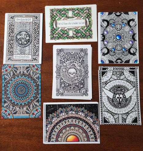 An assortment of A5 size prints on a table; mandalas, celtic knotwork, black ink work, calligraphy