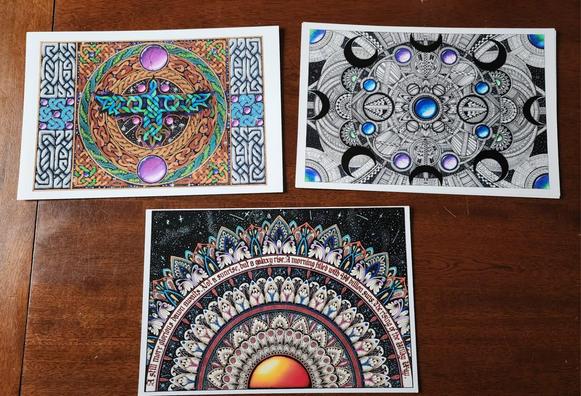 Three A4 size prints on a table; a colourful celtic knotwork piece of a raven, a black ink and colour gemstone intricate geometric pattern, and a half-mandala in colours with some calligraphy inside it