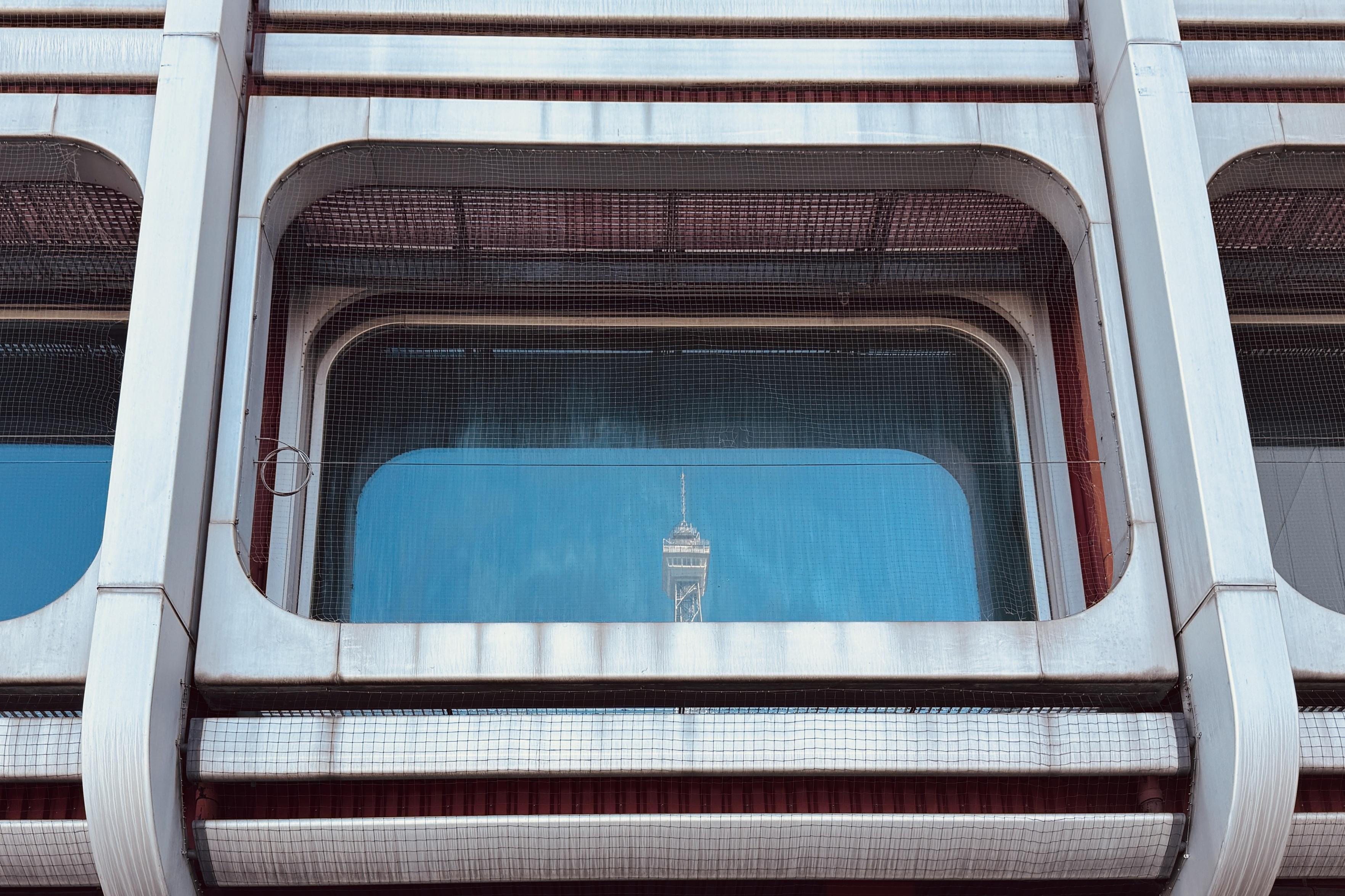 A close-up view of a building's facade featuring modern architectural elements, including large windows with a mesh overlay. The reflection of a tower is visible in one of the windows against a blue sky.
